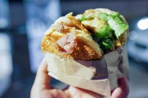 la mano dell'uomo prende una grande pancetta hamburger, tienila e pronta a morderla in bocca, all'evento thailandese foodtruck. foto