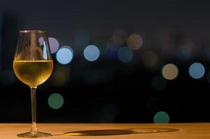 un bicchiere di vino bianco e la sua ombra sul tavolo di legno del bar sul tetto con bokeh colorato di luce della città e spazio per il testo. foto