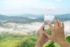 mano che tiene lo smartphone mobile con sfocatura mare di foschia paesaggio mattutino in thailandia foto