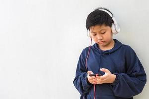 carino ragazzo asiatico con la pelle bianca in una felpa con cappuccio in posa ascoltando la musica su un telefono cellulare e cuffie isolate su sfondo bianco. ritratto e spazio di copia foto