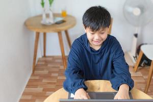 ragazzo asiatico sorridente che chiacchiera con gli amici o uno studente che studia Internet sui social network seduto davanti al laptop a casa. chattare sui social media e videoconferenza. foto