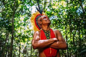 indiano della tribù pataxo, con copricapo di piume. giovane indiano brasiliano che guarda a destra, sorridente e le braccia incrociate foto