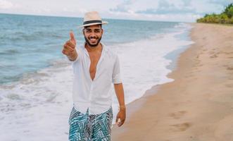 amichevole giovane latinoamericano che fa un gesto con il pollice in su sulla spiaggia, indossando un cappello foto