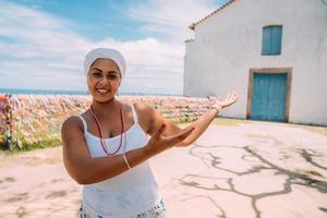 felice donna brasiliana vestita con il tradizionale costume bahiano che mostra qualcosa sul palmo della mano, guarda la telecamera, con il centro storico di porto seguro sullo sfondo foto