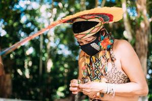 indiano della tribù pataxo con copricapo di piume e maschera protettiva contro la pandemia covid-19. donna indigena brasiliana che fa artigianato foto