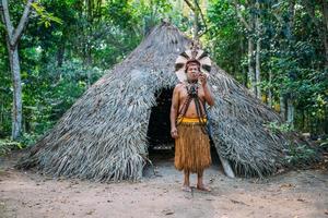 sciamano della tribù pataxo, che indossa un copricapo di piume e fuma la pipa. indiano brasiliano che esamina la macchina fotografica foto