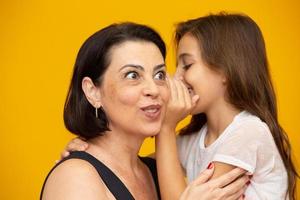 mamma e figlia riservate. bambino che condivide un segreto con la madre. mamma bambino figlia isolata su sfondo giallo ritratto in studio. festa della mamma amore famiglia genitorialità concetto di infanzia foto