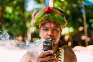 capo indiano della tribù pataxo che fuma la pipa . indiano brasiliano con copricapo di piume e collana che guarda l'obbiettivo. concentrarsi sul tubo foto