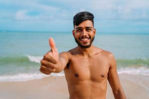 atletico giovane latinoamericano sulla spiaggia con il pollice in su. uomo sorridente che guarda la telecamera foto