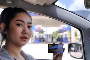 felice bella donna asiatica seduta all'interno della sua auto che mostra il pagamento con carta di credito in una stazione di servizio foto