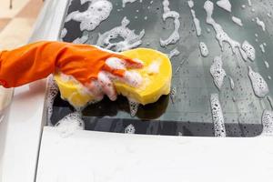 mano della donna che indossa guanti arancioni con spugna gialla per lavare lo specchietto retrovisore auto moderna o pulire l'automobile. concetto di autolavaggio foto