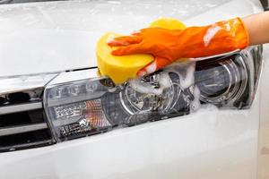 mano della donna che indossa guanti arancioni con spugna gialla che lava il faro di un'auto moderna o di un'automobile di pulizia. concetto di autolavaggio foto