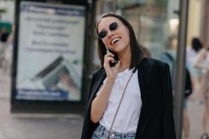 ragazza moderna ed elegante in occhiali da sole, camicia bianca e giacca nera che sorride e parla al telefono su uno sfondo sfocato della città foto