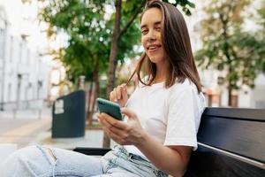 la giovane donna europea felice è seduta per strada sorridendo ampiamente, tenendo in mano lo smartphone e guardando la fotocamera. modello adolescente che riposa all'aperto in città. foto