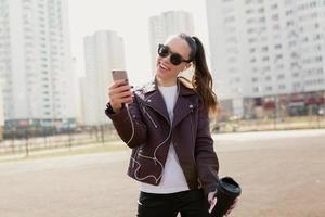 donna alla moda eccitata in giacca e t-shirt bianca divertendosi e usando lo smartphone in città in una calda giornata di sole foto