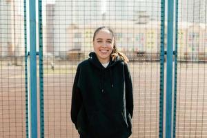 sorridente bella ragazza con i capelli scuri che indossa un pullover nero è in piedi contro il campo sportivo in una calda giornata primaverile e riposa all'aperto foto