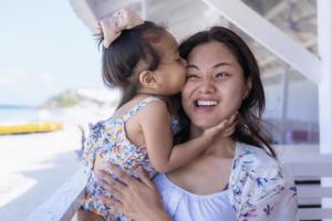 bella giovane madre che ride con una faccia buffa mentre sua figlia le abbraccia il collo e le bacia la guancia all'aperto contro la spiaggia al ristorante o al bar. felice famiglia allegra. madre e bambino che si baciano foto