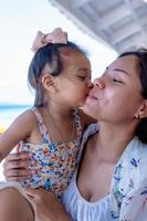 bella giovane madre che ride con una faccia buffa mentre sua figlia le abbraccia il collo e le bacia la guancia all'aperto contro la spiaggia al ristorante o al bar. felice famiglia allegra. madre e bambino che si baciano foto