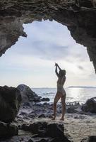 donna magra che alza le mani per lo yoga o posa per aprire la mente alle cose belle. scatto di una donna che pratica yoga mentre si trova in una grotta con vista sul mare all'alba o al tramonto. foto