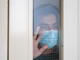 uomo solo in maschera medica che guarda attraverso la finestra. isolamento domiciliare per auto quarantena. concetto di quarantena domestica, prevenzione covid-19. situazione dell'epidemia di coronavirus foto