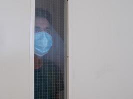 uomo solo in maschera medica che guarda attraverso la finestra. isolamento domiciliare per auto quarantena. concetto di quarantena domestica, prevenzione covid-19. situazione dell'epidemia di coronavirus foto