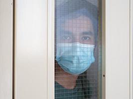 uomo solo in maschera medica che guarda attraverso la finestra. isolamento domiciliare per auto quarantena. concetto di quarantena domestica, prevenzione covid-19. situazione dell'epidemia di coronavirus foto