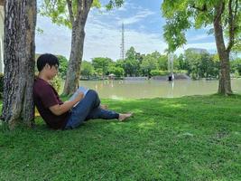 il giovane uomo asiatico con gli occhiali con i capelli neri si siede sotto un albero con in mano un libro. felice e rilassati. cosa stai pensando nella città del parco estivo foto