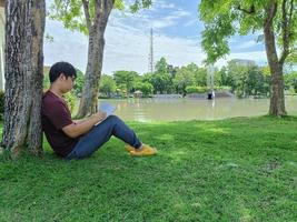 il giovane uomo asiatico con i capelli neri si siede sotto un albero con in mano un libro. stress e ansia. cosa stai pensando nella città del parco estivo foto