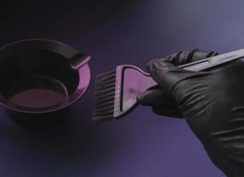 una mano guantata tiene una spazzola per tinture per capelli. sfondo futuristico parrucchiere in luce al neon viola, modello di parrucchiere alla moda. foto