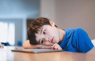 bambino della scuola che usa il tablet per i compiti, viso annoiato del bambino sdraiato a testa in giù guardando fuori nel profondo dei suoi pensieri, ragazzo che impara l'aula online a casa, concetto di e-learning o homeschooling foto