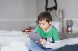 ragazzo che usa il tablet che gioca su internet con un amico, bambino che fa i compiti online con il pad digitale a casa, bambino seduto sul letto a rilassarsi, guardare cartoni animati o parlare online con un amico foto