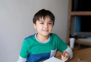 ragazzo felice che usa il disegno a matita o disegna su carta, bambino carino che guarda la fotocamera con la faccia sorridente che si siede sul tavolo a fare i compiti, il bambino si diverte con l'arte e l'attività artigianale a casa, concetto di educazione foto