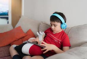 felice ragazzo che indossa le cuffie per giocare su internet con gli amici, bambino seduto sul divano a leggere o guardare cartoni animati sul tablet, bambino a scuola che fa i compiti online a casa. foto