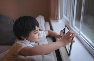 ritratto cinematografico un ragazzo felice che gioca a un gioco online sul tablet, bambino che guarda cartoni animati da internet, bambino seduto sul divano accanto alla finestra con una giornata piovosa al mattino foto