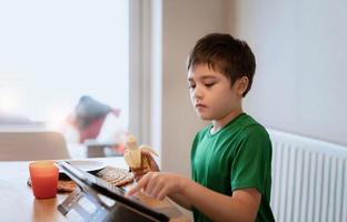 bambino che mangia banana e guarda cartoni animati sul tablet, bambino che fa colazione e gioca online su internet con gli amici prima di andare a scuola la mattina foto