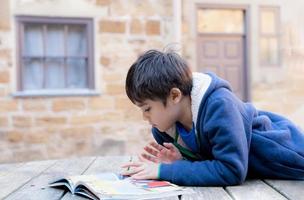 bambino della scuola che fa il libro di lettura dei compiti, giovane ragazzo del ritratto seduto da solo fuori a leggere una storia, concetto di educazione foto