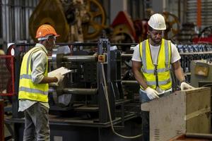 il team di un lavoratore industriale afroamericano sta controllando il valore di configurazione della macchina per la formatura di rotoli di lamiera tramite un monitor automatizzato mentre lavora all'interno della fabbrica di tetti per l'industria della sicurezza