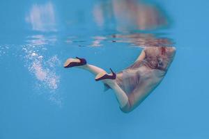 ritratto d'arte surreale di giovane donna in abito grigio, sciarpa di perline, piccola borsa, tacchi alti viola sott'acqua in piscina foto