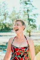 donna bionda emotiva che ride con i capelli bagnati che fanno schizzi d'acqua. vacanze, felicità, divertimento, estate, concetto di svago foto