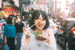 giovane donna asiatica adulta che mangia cibo di calamari alla griglia speziati al mercato di Chinatown del sud-est asiatico foto