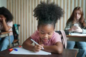 bambino della ragazza in età prescolare che disegna con la matita di colore su carta bianca sul tavolo in classe con gli amici foto