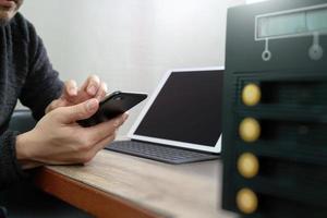 Mano dell'uomo d'affari tramite smartphone, pagamenti mobili acquisti online, canale omni, computer con tastiera docking per tablet digitale, server compatto sulla scrivania in legno foto