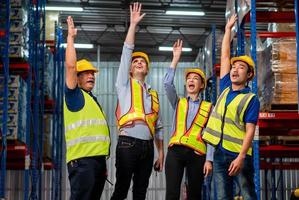 squadra di magazzinieri di successo che prendono a pugni l'aria per festeggiare, lavoratori manuali che celebrano il successo nel magazzino della fabbrica, lavoratori con elmetto protettivo per festeggiare sul posto di lavoro foto