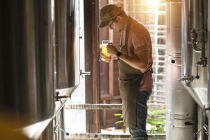 un giovane lavora in un birrificio e controlla la qualità della birra artigianale. il proprietario del birrificio assaggia le migliori birre di bach. la scorciatoia di un uomo riempie un bicchiere di birra foto