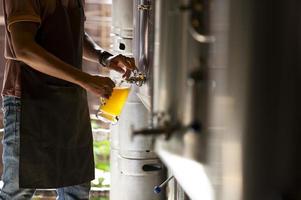 un giovane lavora in un birrificio e controlla la qualità della birra artigianale. il proprietario del birrificio assaggia le migliori birre di bach. la scorciatoia di un uomo riempie un bicchiere di birra foto