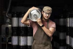 giovane uomo in grembiule di pelle che tiene un fusto di birra al moderno birrificio, operaio del birrificio artigianale foto