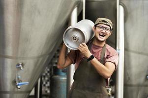 giovane uomo in grembiule di pelle che tiene un fusto di birra al moderno birrificio, operaio del birrificio artigianale foto