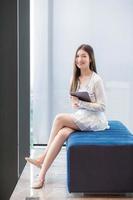 la giovane donna d'affari asiatica professionale con i capelli lunghi in camicia bianca tiene il tablet sorridendo felicemente mentre si siede sulla sedia nell'ufficio del posto di lavoro. foto