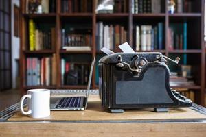 vecchia macchina da scrivere vintage ricoperta di polvere con foglio di carta bianca vicino a un taccuino moderno e una tazza di caffè sullo sfondo della libreria. tecnologia moderna ed elettrodomestici vintage foto