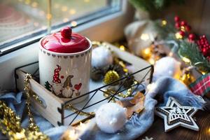 decorazioni natalizie vicino alla finestra su un accogliente davanzale in legno con una tazza con un drink e un barattolo di biscotti. lucine, ramo di albero di natale, vassoio per la colazione natura morta foto